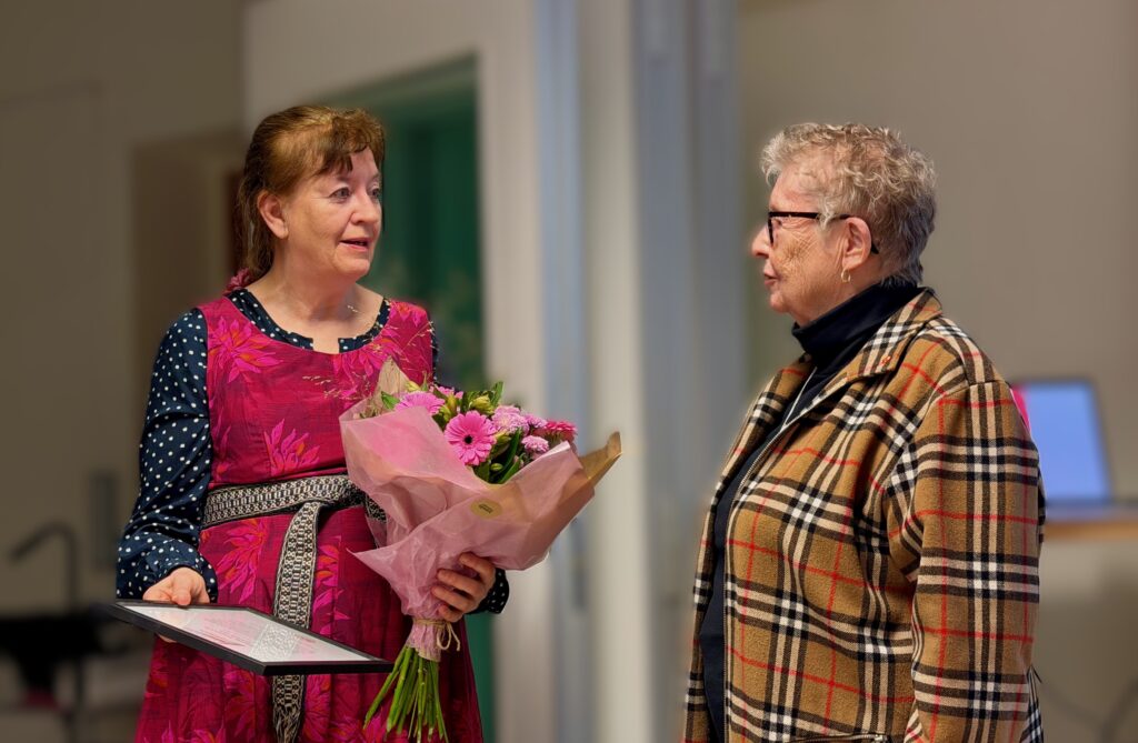 Två kvinnor talar med varandra. Den ena håller en blombukett och ett diplom i händerna.